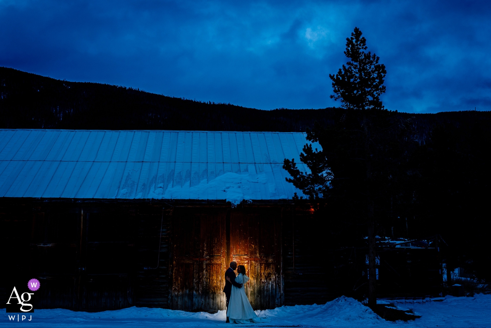 Foto von: <a href="https://www.wpja.com/photos/artistic-wedding-photographers-portrait/2343896"> Trent Gillespie - Silverthorne Colorado Colorado-Hochzeitstagbraut- und -bräutigamporträt im Schnee an der Dämmerung gegen eine beleuchtete Scheune