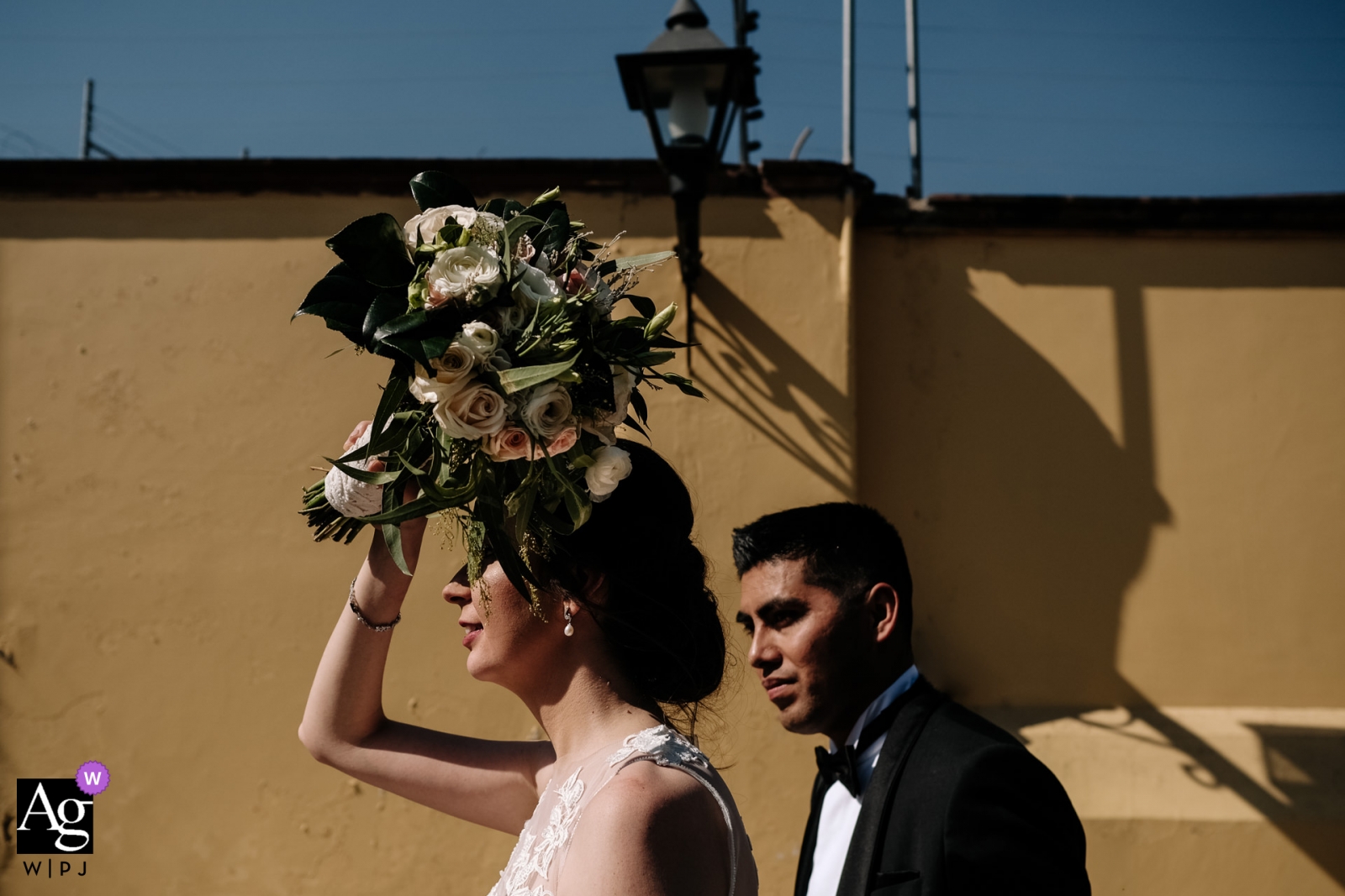 Ein Porträt eines fröhlichen, frisch verheirateten Paares, das in Oaxaca-Stadt, Mexiko, in der Sonne spaziert, wobei die Braut ihr Gesicht mit ihrem Blumenstrauß vor der Sonne schützt