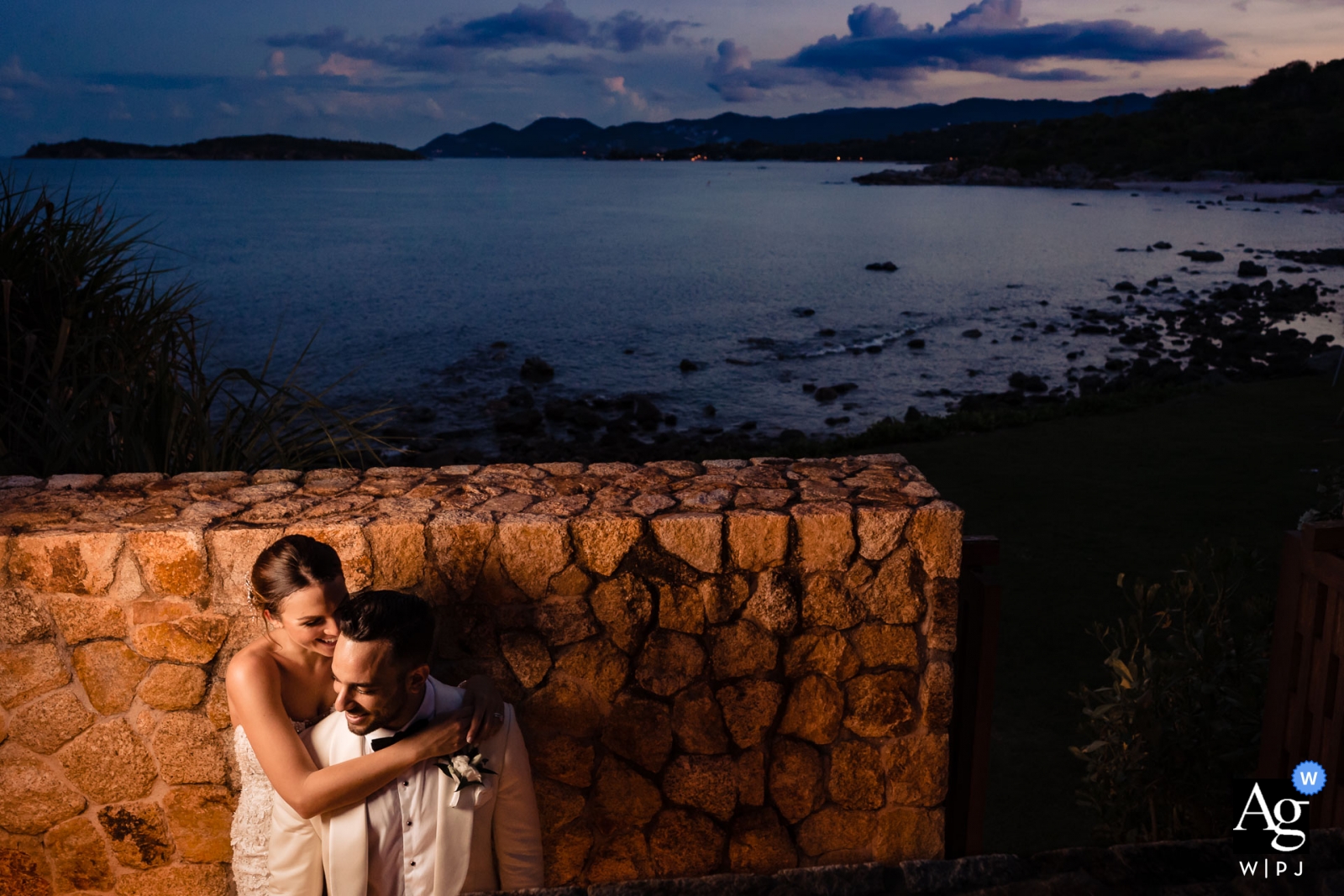 Una imagen romántica de una pareja abrazándose al atardecer frente a un muro de piedra con vista al agua en Samujana, Koh Samui, captura la esencia del día de la boda.