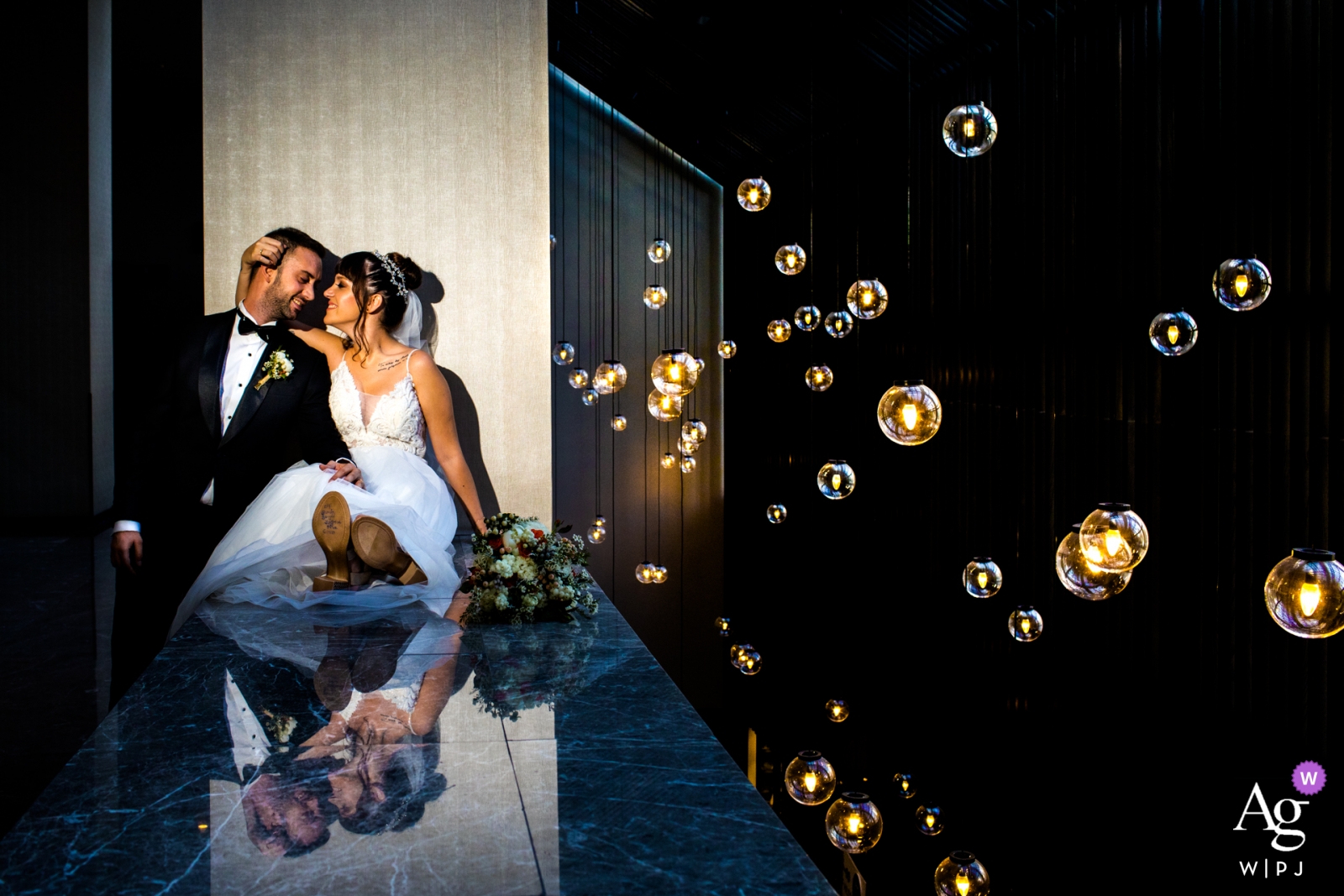 Bride is sitting on a platform and groom is next to her close to gallery space with lamps hanging from the ceiling at the Hilton Ankara