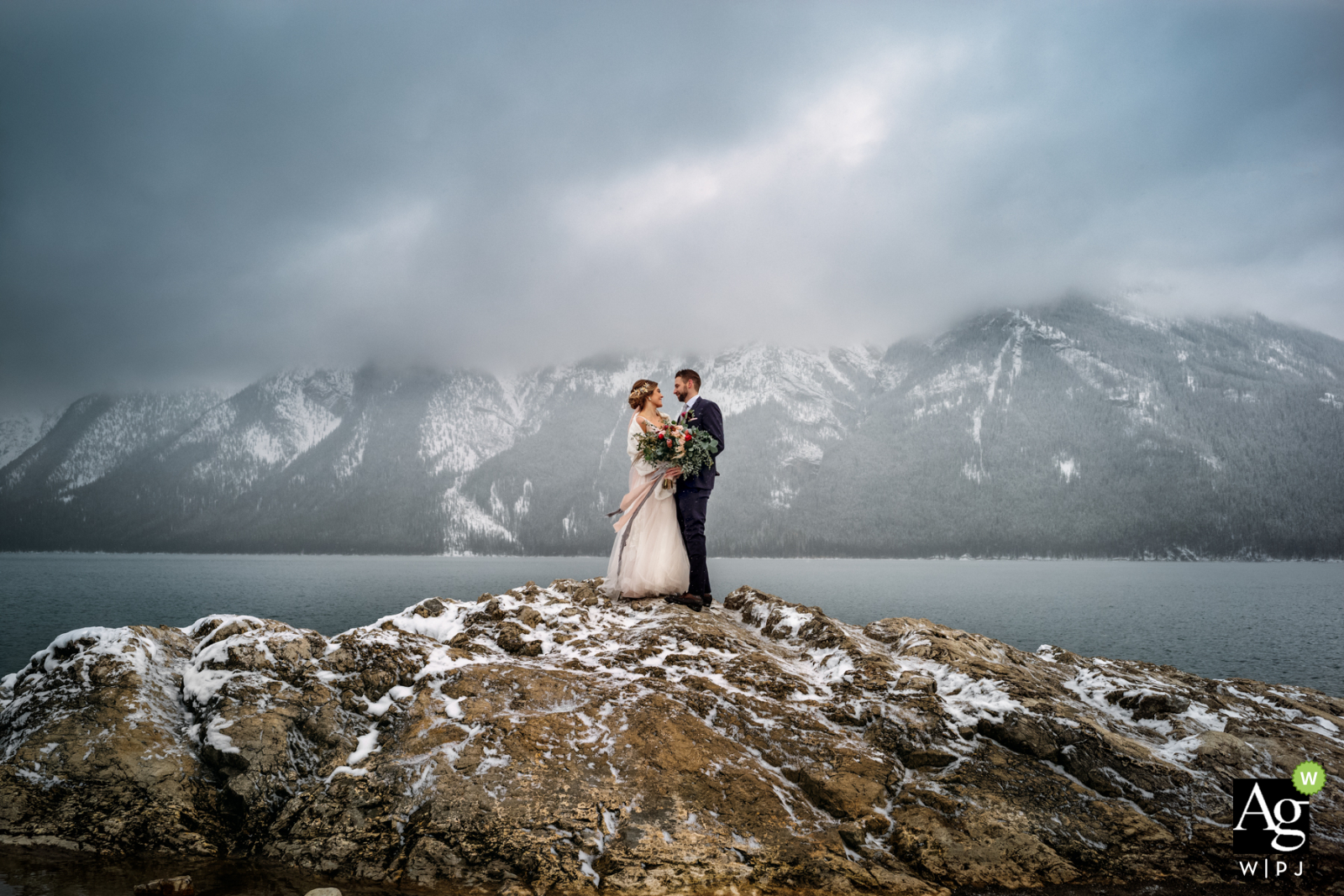 Lago Minnewanka, Parque Nacional de Banff, AB, Canadá casal de noivos posou sessão de retratos da noiva e do noivo abraçados entre as montanhas cobertas de neve