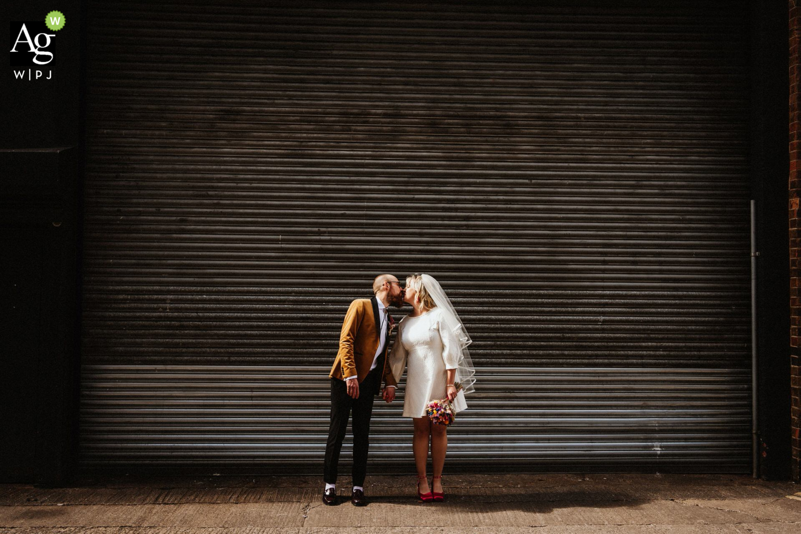 The couple shared a brief kiss shoulder width apart in front of a large corrugated metal door at Trafalgar Warehouse in Sheffield, UK, with the sun streaming through the gaps in the building and creating a warm, romantic atmosphere