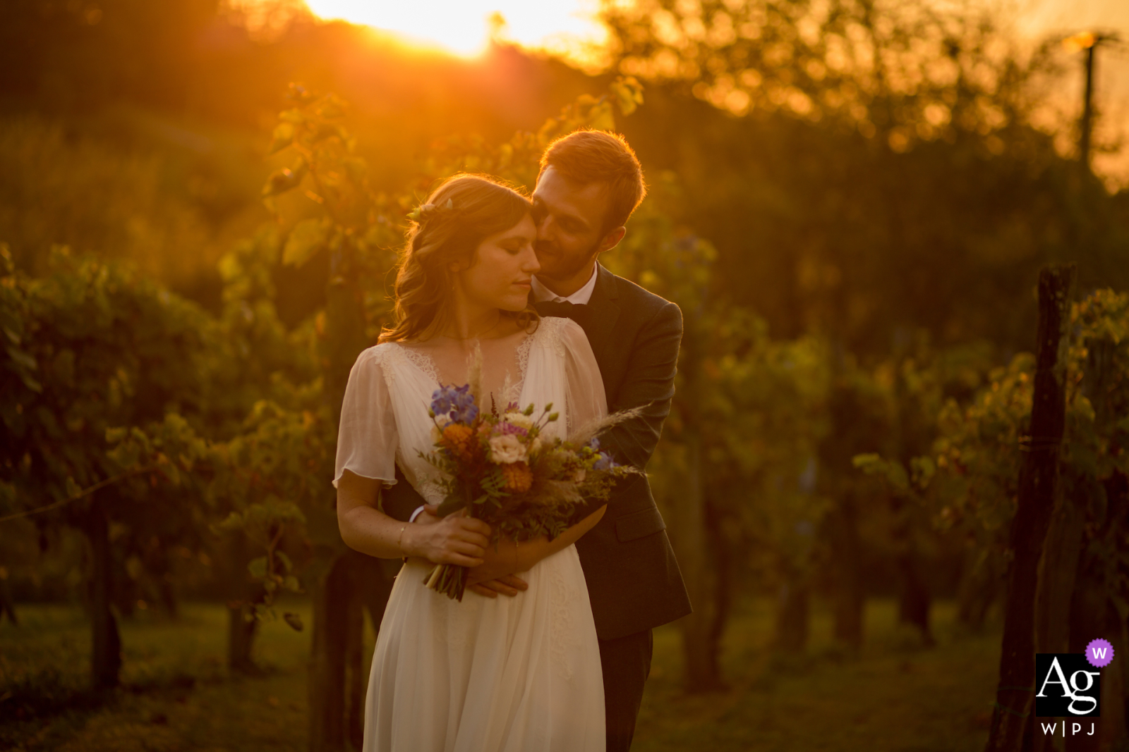 The bride and groom are embracing in the breathtaking sunset of the French countryside, with the bride clutching a bouquet, at a vineyard in Nouvelle-Aquitaine at their wedding reception