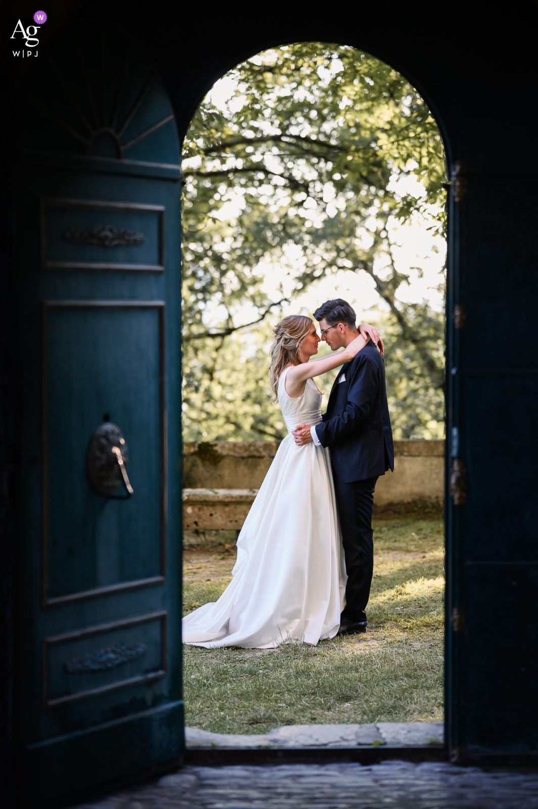 Couple in wedding attire kisses in front of Château de Goudourville, with the castle gate in the foreground creating a picturesque frame for their affectionate embrace