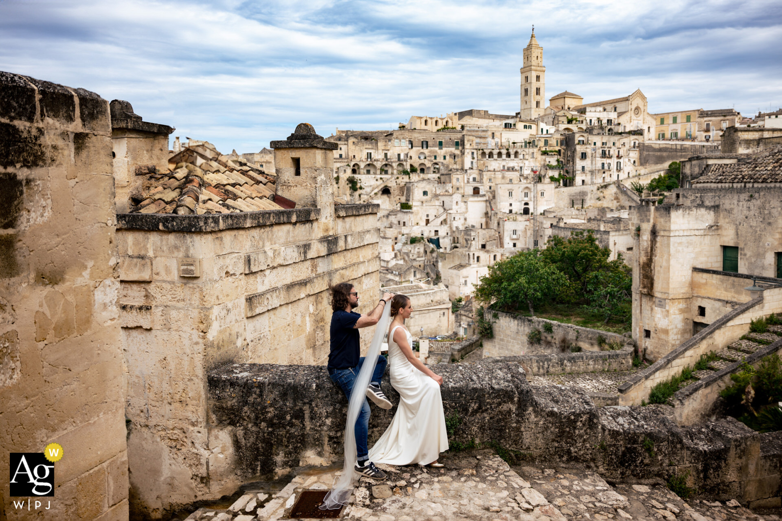 Matera: Bride perched on a stone wall overlooking village, receives assistance in fixing veil