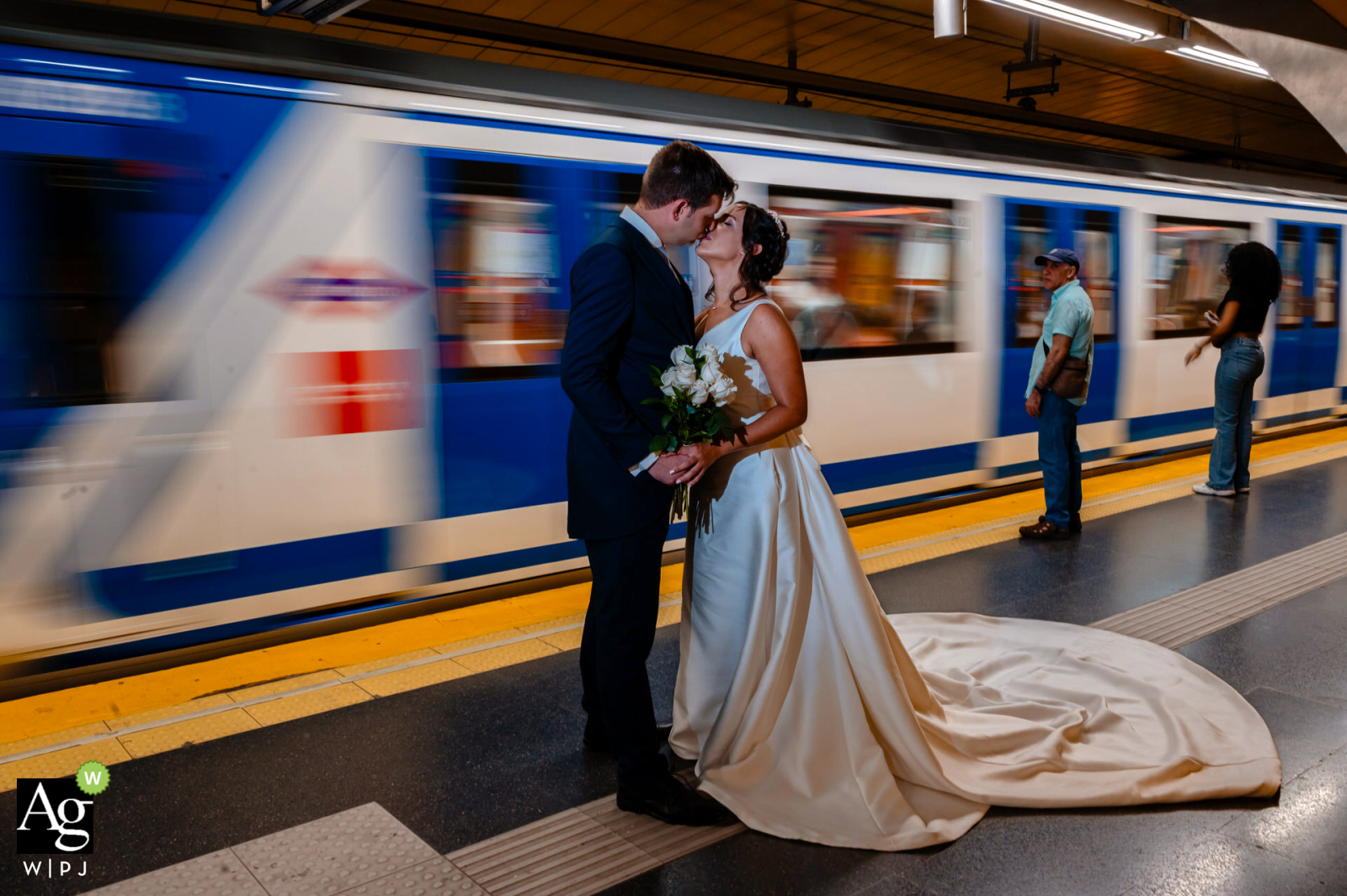 In an urban setting in Madrid, Spain, a couple in their wedding attire hugs each other tightly while standing beside a swiftly moving subway train, creating a dynamic and romantic scene.
