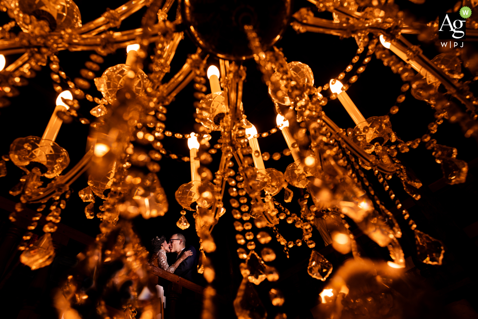 A stunning wedding photo captured at Domaine de la Poudié, Tarn, France. The couple is elegantly framed through a chandelier on a balcony, creating a beautiful and intimate portrait.