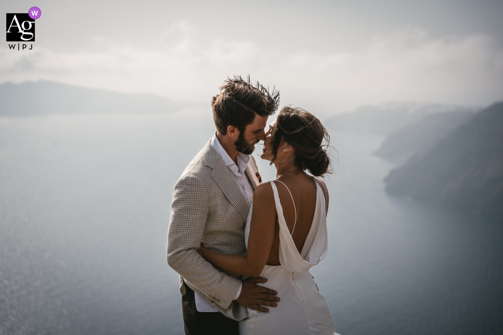 In Santorini, Greece, a bride and groom share a romantic kiss against the backdrop of the clear sea. The photographer captures their love from a high viewpoint, creating a dreamy image.