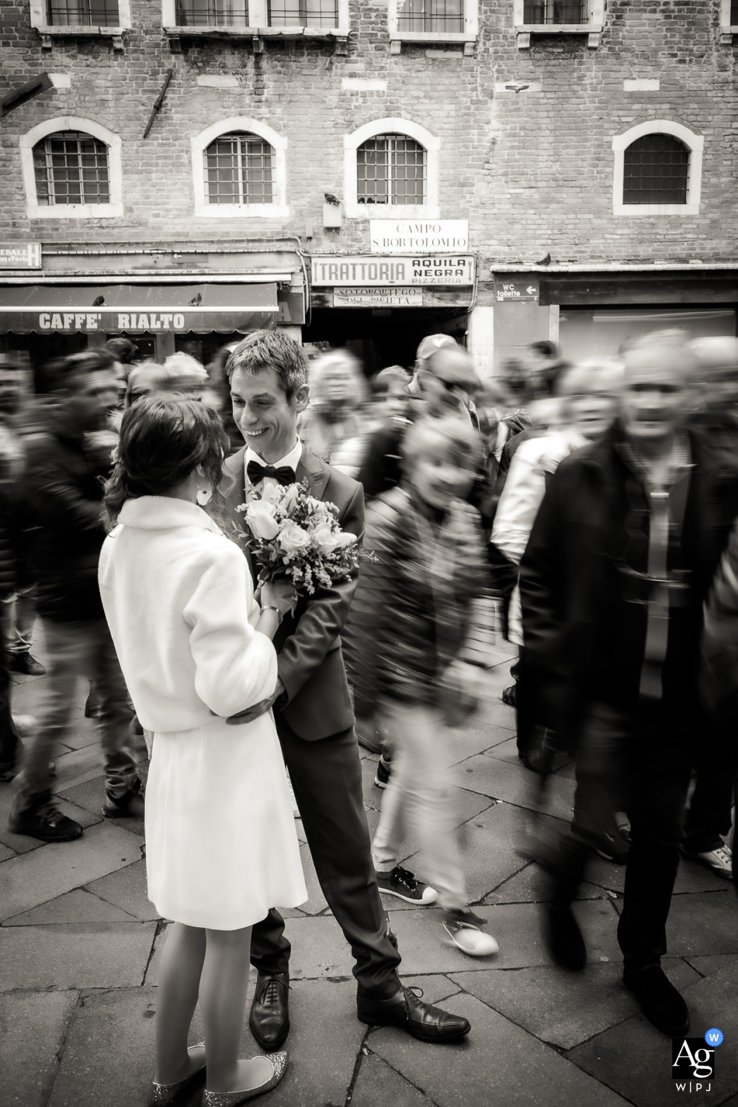 A talented wedding photographer captured a beautiful portrait of a bride and groom at Campo San Bortolo in Venice, Italy. The image showcases the romantic atmosphere as the couple stands still while passersby blur in the background.