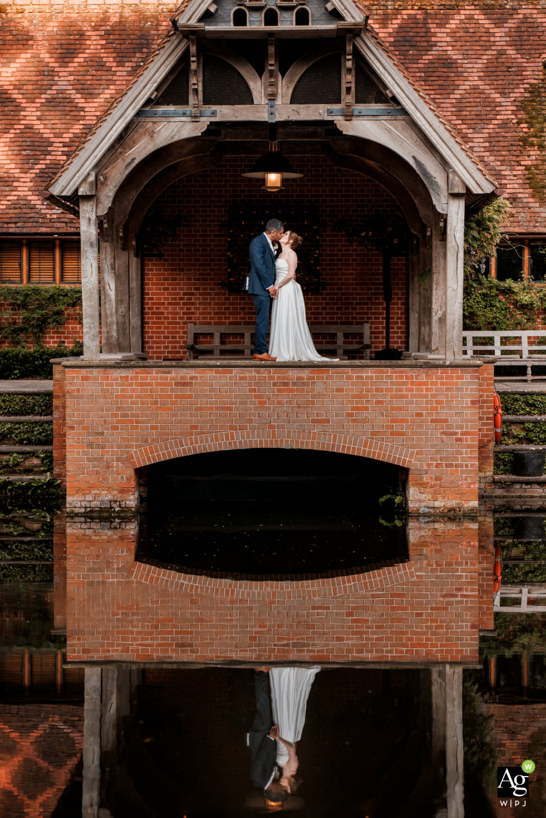 At The Dairy in Buckinghamshire, UK, a long shot captures the bride and groom standing near the lake. Their reflection shimmers on the water's surface, creating a serene and picturesque scene that highlights the natural beauty of their venue building.