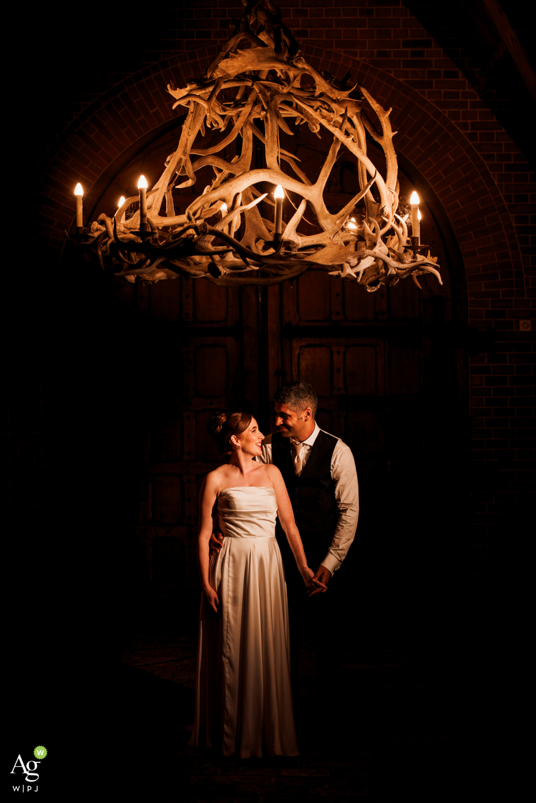 At The Dairy in Buckinghamshire, UK, the bride and groom stand together under soft, evening light. A warm antler chandelier glows above them in a vertical frame, adding an elegant and rustic charm to their romantic portrait.