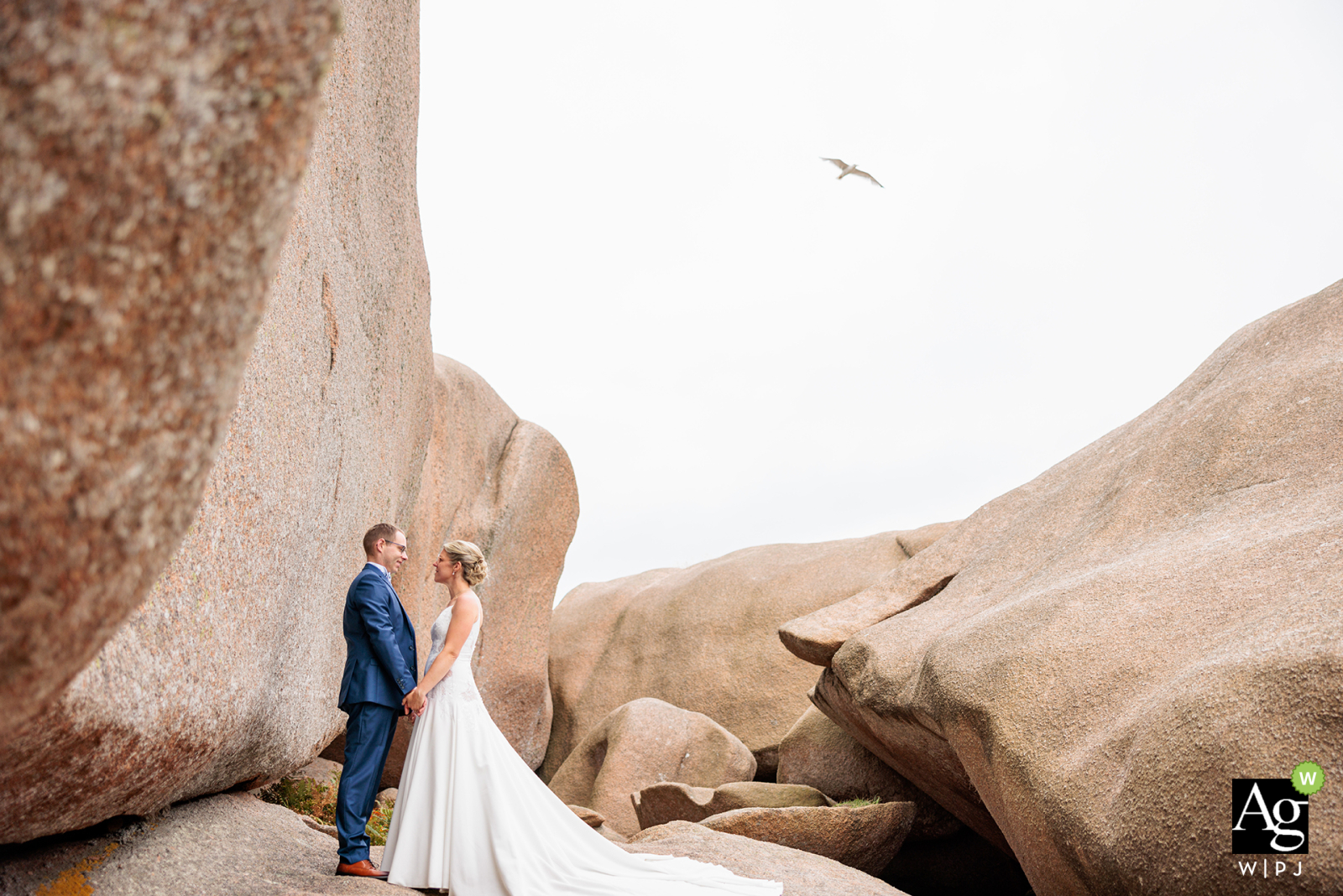 Bride and groom pose against a dramatic sand-colored cliff and boulder backdrop in Tregastel, France. The couple embraces amidst the rugged landscape in a scenic wedding portrait.