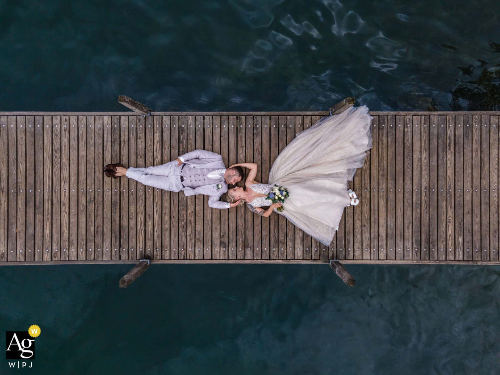 A tranquil lakeside drone portrait captured by a skilled WPJA wedding photographer. See the bride and groom from a unique angle on a wooden dock in the stunning Lake Of Zoug, Switzerland.
