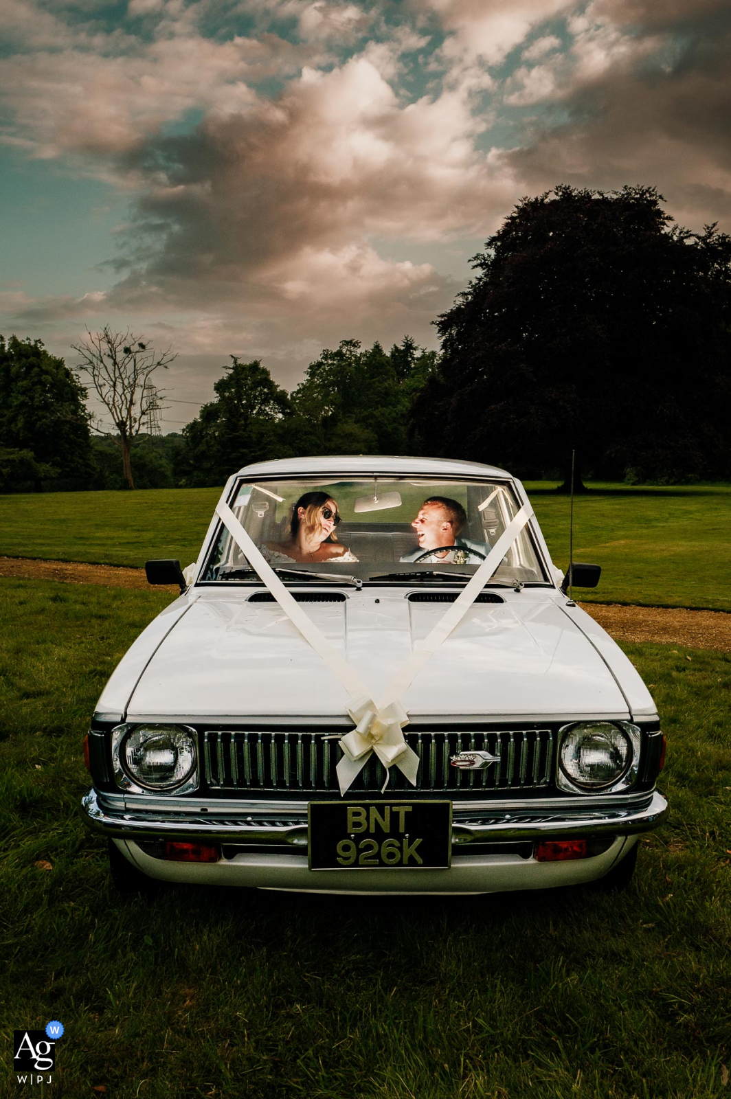 At Hale Park House, Dorset, UK, the couple sits inside the groom's vintage car. A flash under the dashboard highlights their spontaneity, framed by a beautiful sunset.