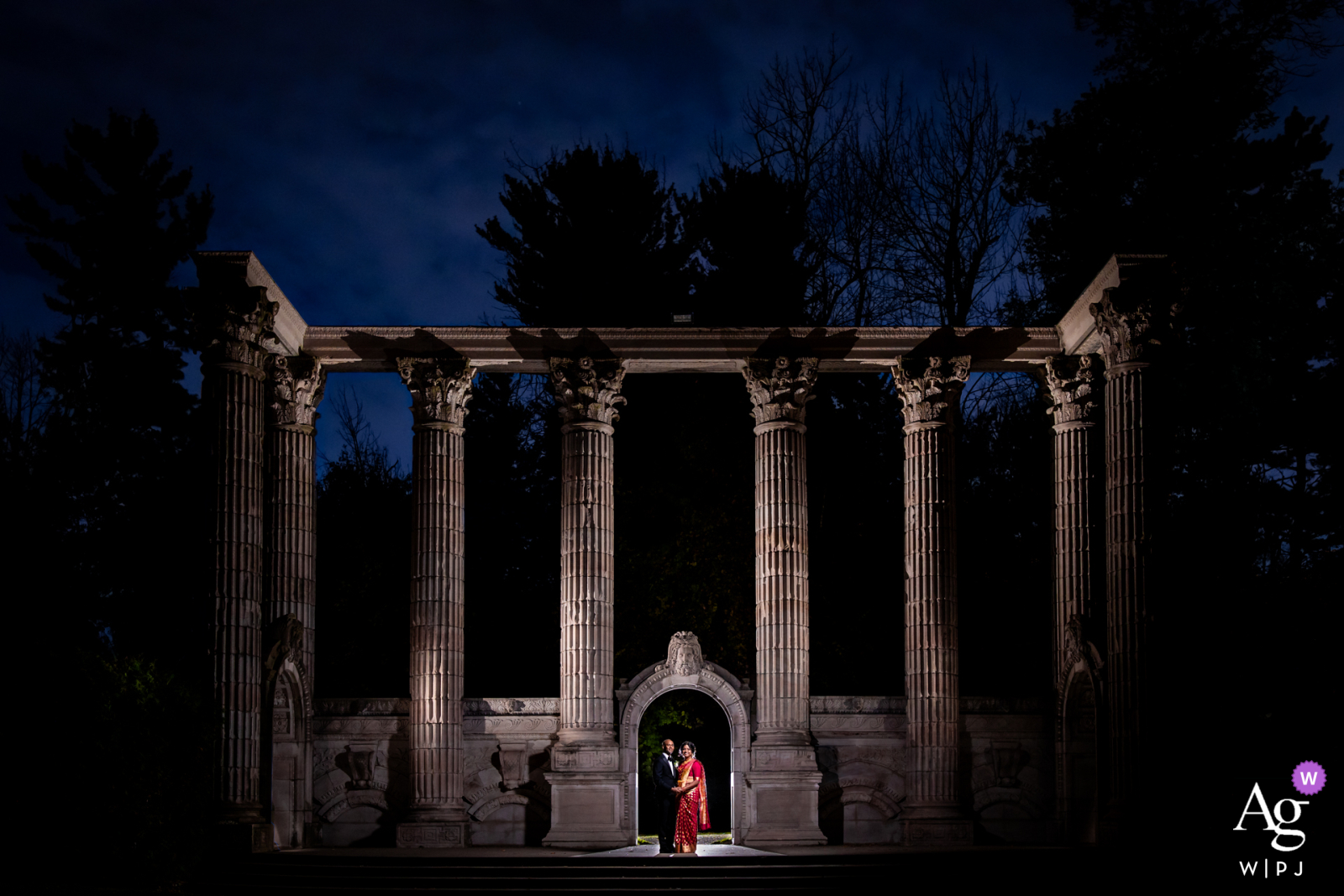In Toronto, Canada, a night-time photo beautifully frames a couple in a stone archway, surrounded by grand stone pillars.
