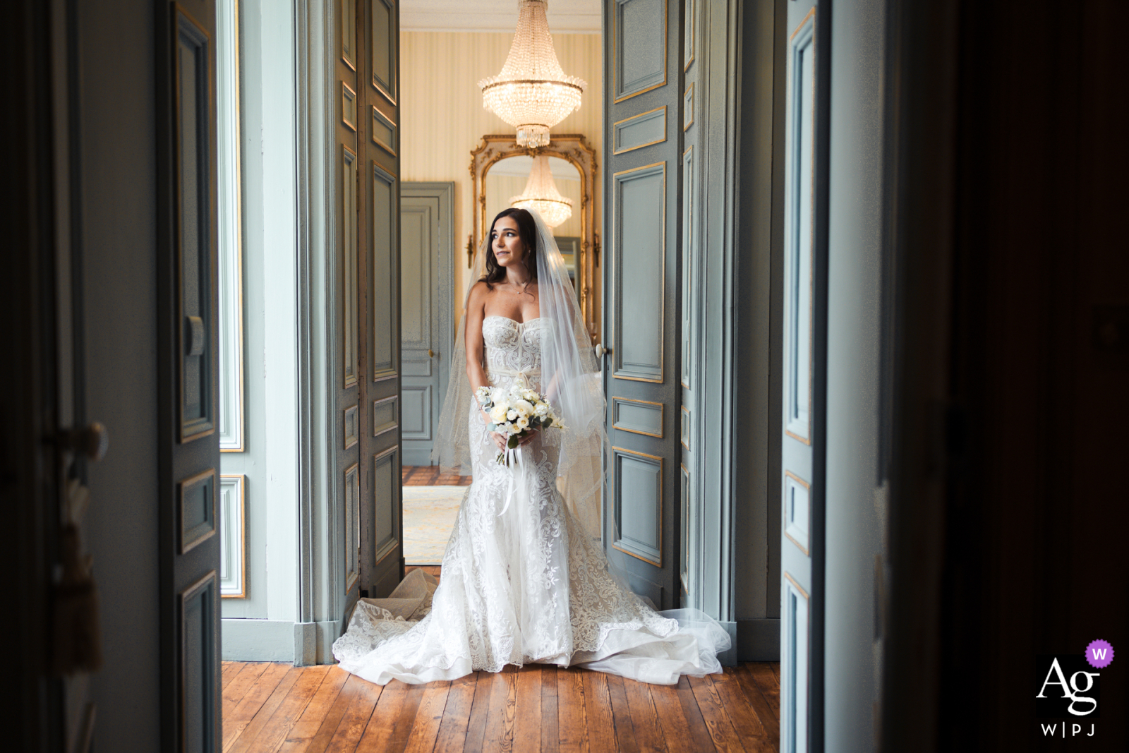 At Chateau La Durantie in France, the bride gracefully poses inside the castle, standing at the center of a hallway. She gazes off to the side as soft window light bathes the scene, highlighting her elegance and the timeless beauty of the setting.