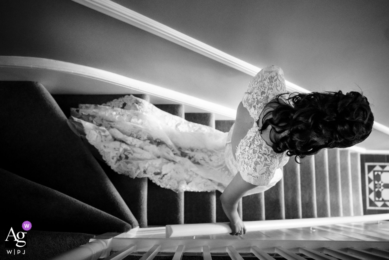 At the bride's home in Hertfordshire, a black-and-white top-down view captures the bride as she descends the stairs to see her family, highlighting the intricate lace details on the back of her dress.