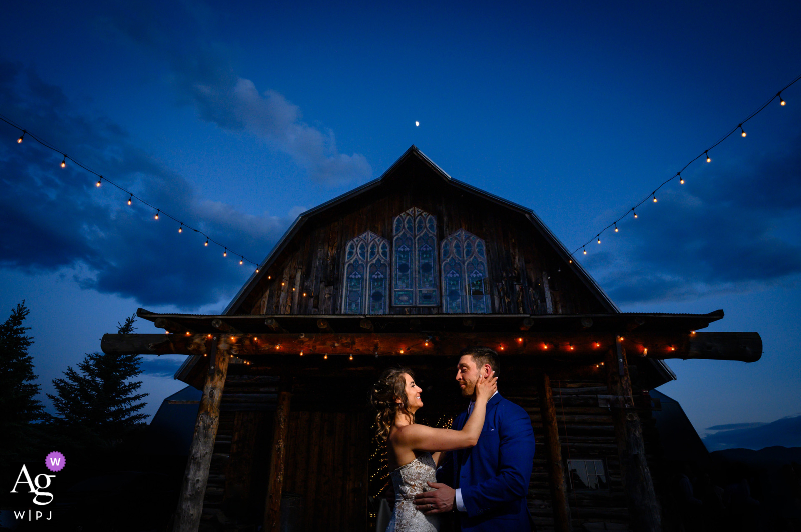 At Evergreen Memorial Barn in Evergreen, Colorado, the couple shares a laugh in front of their wedding reception venue at dusk, with the moon shining brightly overhead, adding a romantic and serene backdrop.