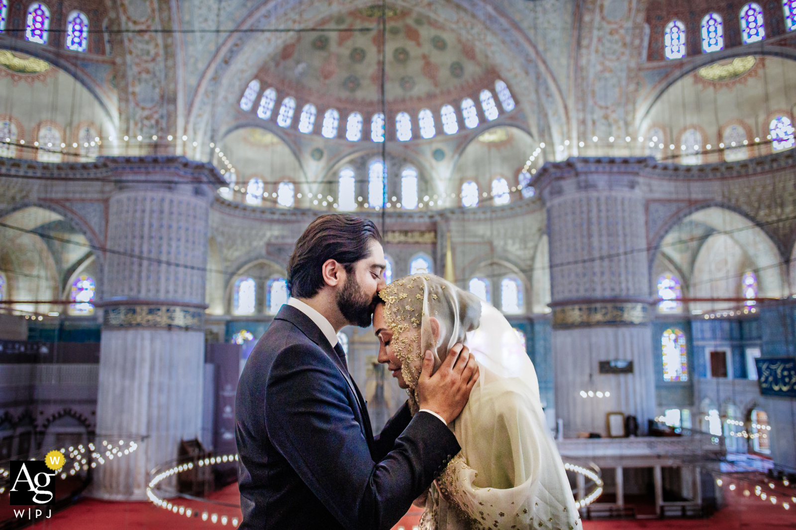 Blue Mosque, Istanbul, Türkiye — In a symmetrically framed wedding portrait, the groom kisses the bride’s head as they stand in profile, with the historic arches and domes of the Blue Mosque providing a dramatic old-world backdrop.