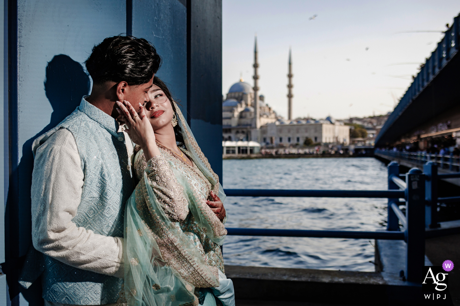 Galata Bridge, Karakoy, Istanbul — The couple poses for a wedding day portrait on the dark blue-painted bridge, dramatically lit by the photographer’s off-camera flash against the city and water backdrop.