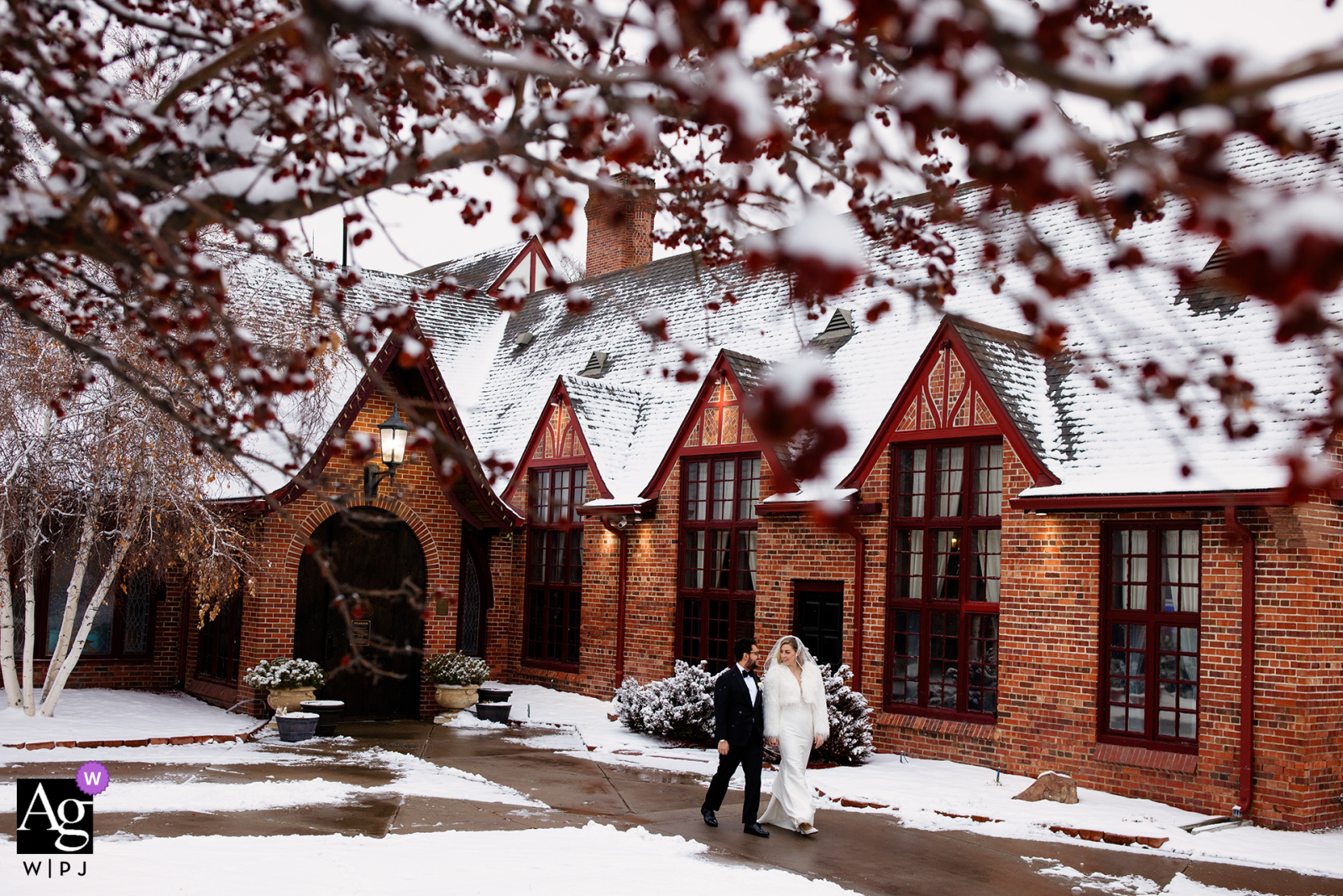The bride and groom walk through the snowy courtyard at Wellshire Event Center, their figures framed by the softly illuminated architecture and snow-dusted trees, creating an artistic scene-setter of their Colorado wedding day.