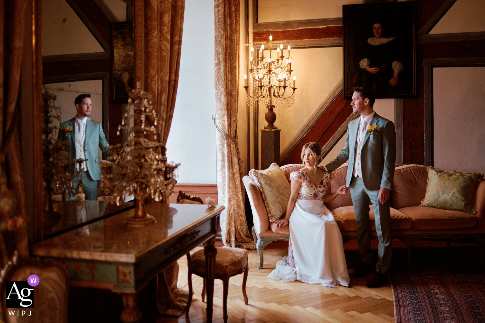 Fribourg, Switzerland — Indoors at the ceremony location, the bride sits on a couch in the magnificent castle lounge while the groom stands behind her with his hand on her shoulder, his reflection visible in the mirror for an elegant wedding portrait.