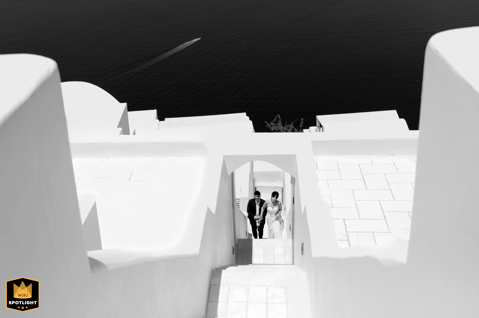 Newlyweds walk up immense white stairs at Andromedas Villa & SPA Resort in Santorini, Greece. Black and white shot emphasizes the contrast against the stunning sea backdrop.