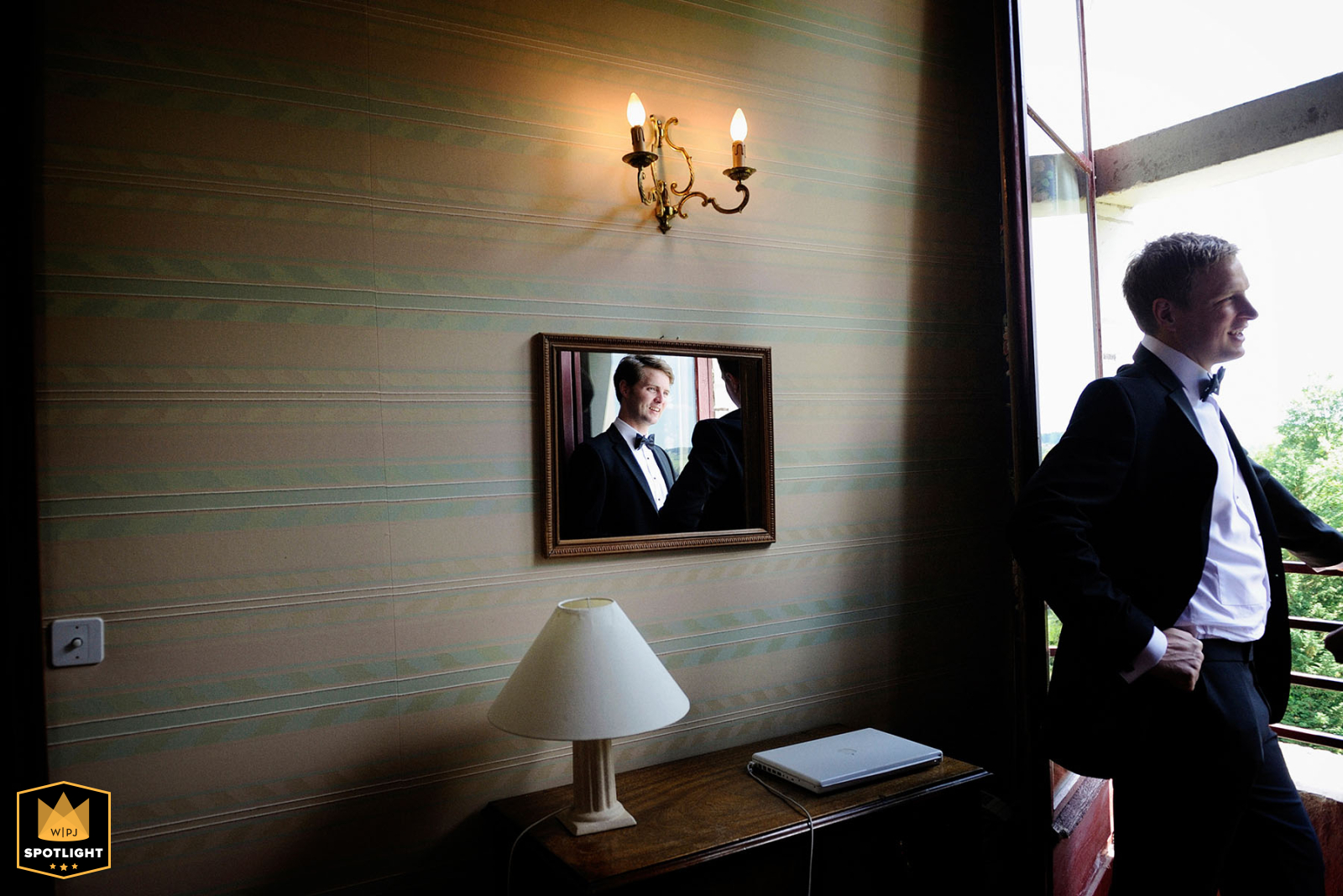 Groom and best man sharing a relaxed moment by the hotel window at Chateau de Chissay in Touraine, France, with a reflection visible in the wall mirror.