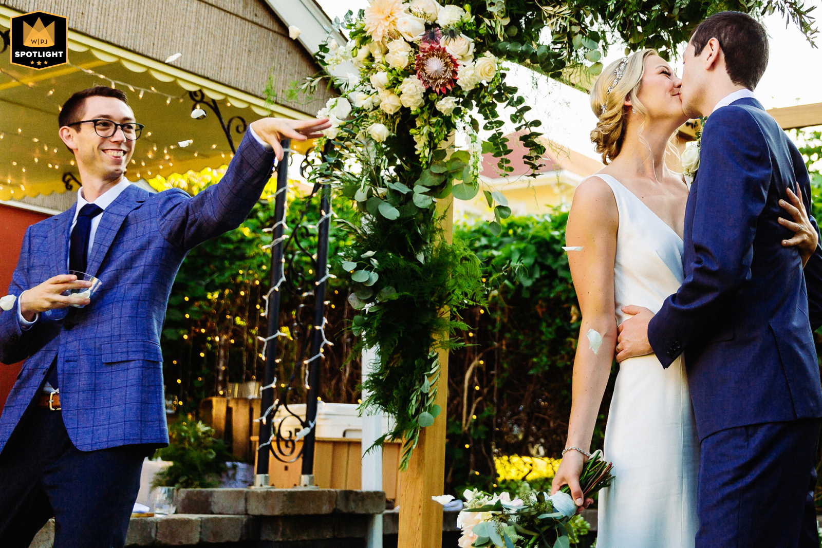 Un momento conmovedor capturado en una boda en el patio trasero de Watertown, Massachusetts, cuando el hermano de la novia fotografía con alegría el beso romántico de la pareja, agregando un toque de diversión a la imagen de la boda.