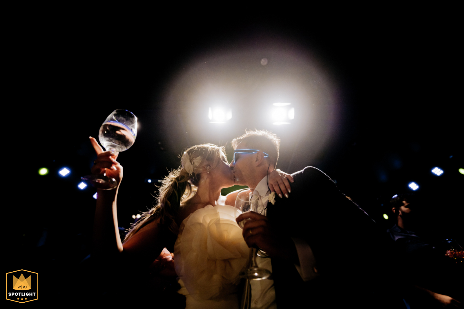 A romantic scene captured at a wedding in São Caetano do Sul, São Paulo. In a backlit frame against a black background, the couple shares a kiss as they raise their glasses in celebration.