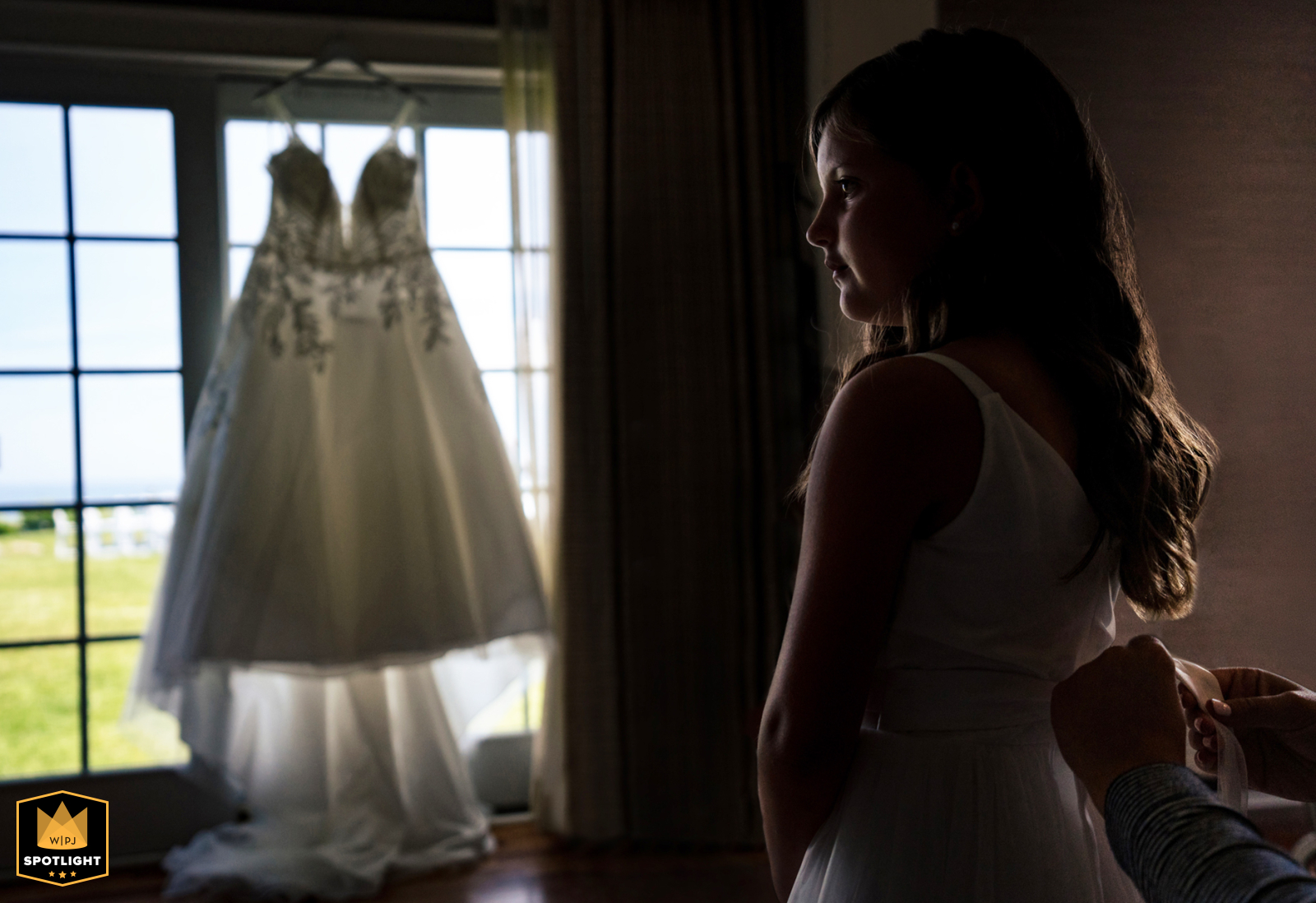In York, Maine, the flower girl dresses, with the bride's gown hanging in the background. This scene captures the anticipation and excitement of getting ready, highlighting the charming contrast between the little girl's preparation and the gown.