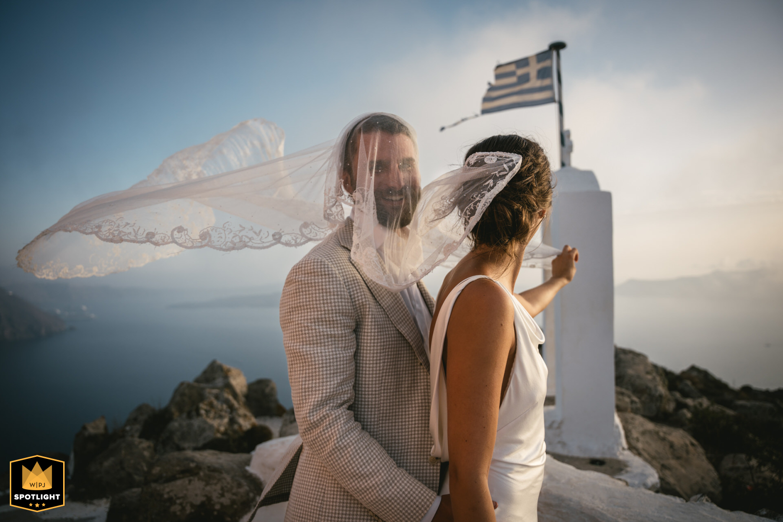 A groom in Santorini, Greece, is momentarily entangled in his bride's veil. The wedding photo captures the candid moment.
