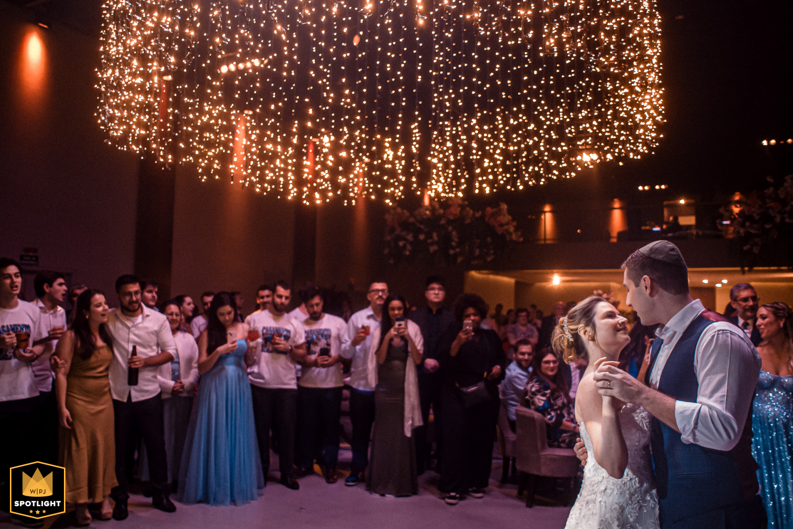 At Espaço Welucci – São Paulo, Brasil, the bride and groom share their first dance, positioned to the far right, with a row of guests filling the background.