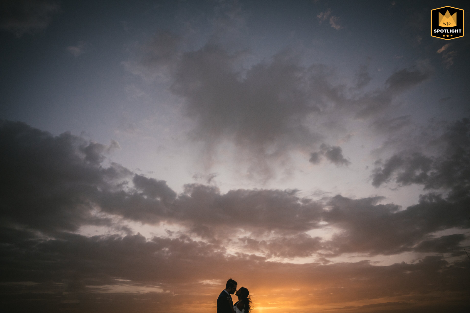 Silhouetted Couple Poses Low on the Horizon Against a Dramatic Sunset Sky in Portugal A couple is silhouetted against a sunset in Portugal. They pose low to the horizon with a big sky above, creating a dramatic and romantic portrait.