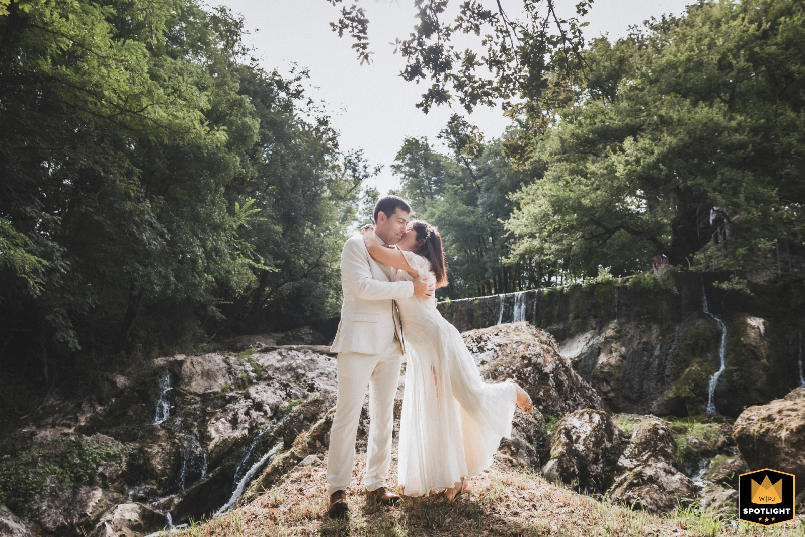 The bride and groom embrace in front of a pretty waterfall in Salies-de-Béarn, France. This intimate scene is captured next to the family home, a romantic setting for their union.