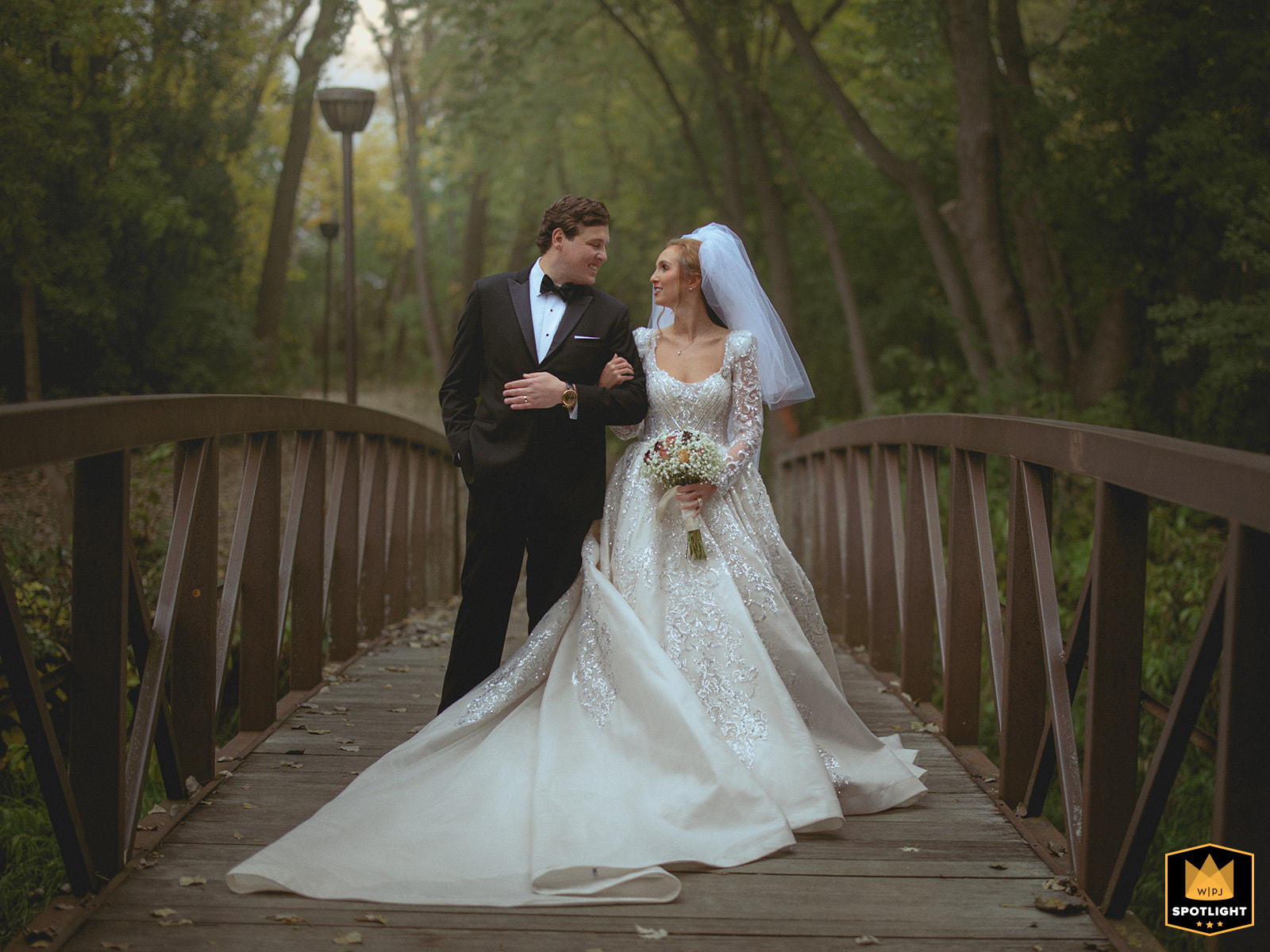 The bride and groom walk arm in arm across a wooden bridge at Gaelic Park. They smile at each other, captured in a serene forest setting during their wedding.