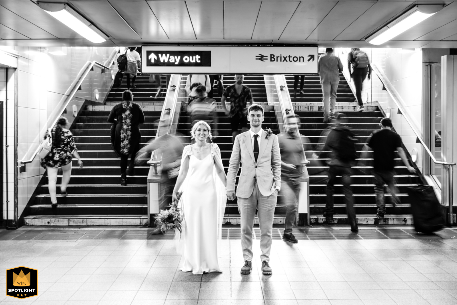 Couple stands intimately in Brixton Tube Station, a blur of commuters and London's vibrant life surrounding them.