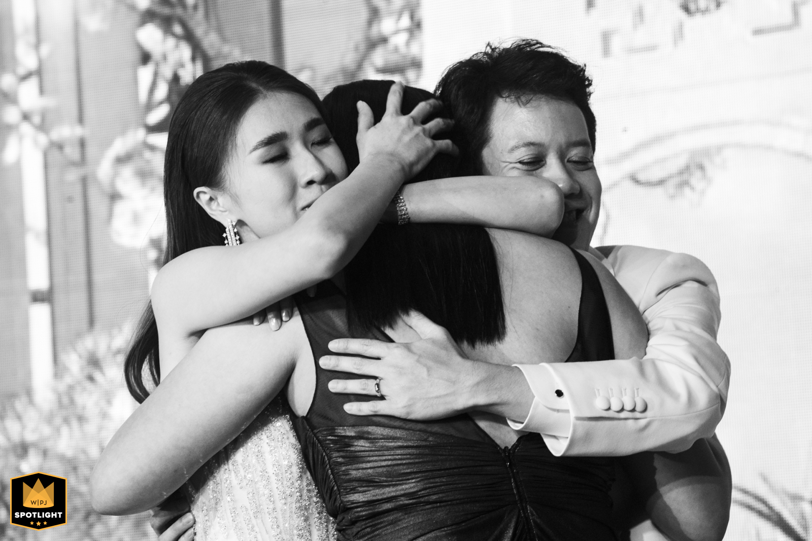 A black-and-white image at Four Seasons Bangkok shows a couple warmly hugging a woman, all three sharing an emotional embrace that expresses affection and heartfelt connection during the celebration.