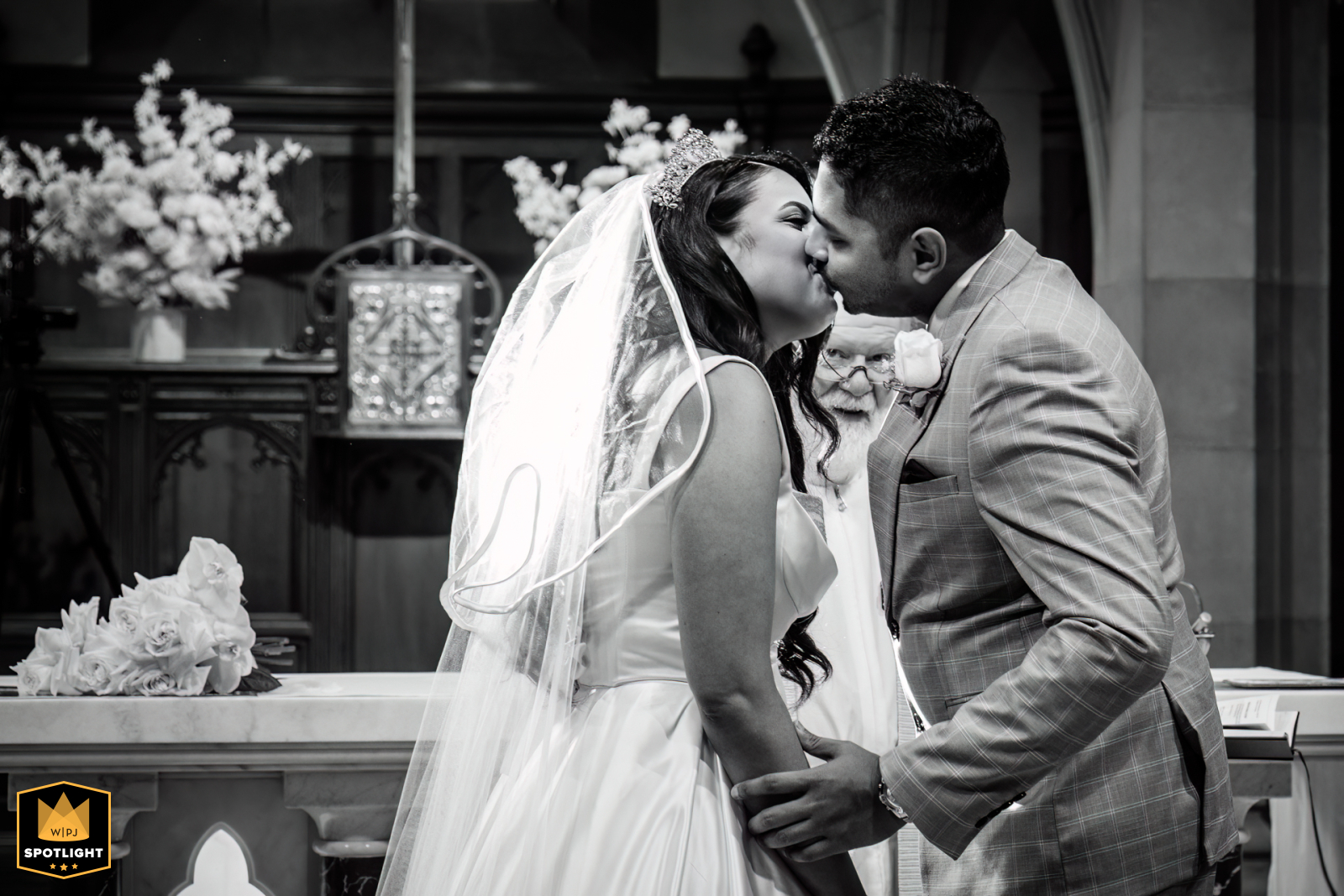 Inside a Melbourne church in Victoria, Australia, a black and white image shows the priest standing behind the couple, discreetly watching as they share their first kiss, adding a lighthearted and candid touch to the ceremony.