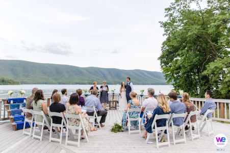 A couple exchanges vows on a deck overlooking a serene mountain lake, surrounded by loved ones.