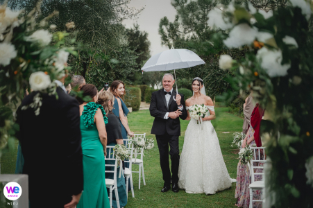 Father and daughter walk hand in hand toward the altar, surrounded by gentle rain at Rocca di Cetona in Tuscany, Italy. The lush landscape frames their heartfelt journey on this intimate wedding day.