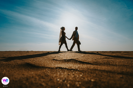 Couple walks hand in hand on a sunlit shore, silhouettes contrasting with the bright sky.
