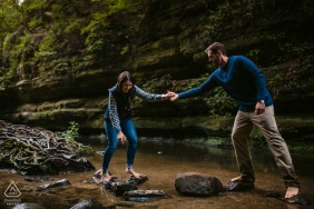 Starved Rock State Park Fine Art Engagement Image in Illinois as The couple is helping each other over a rocky stream while barefoot