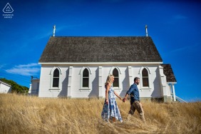 A couple strolls hand in hand through a sun-drenched field of dry grass, with a quaint white church in the background, during their Tiburon, CA engagement portrait session.