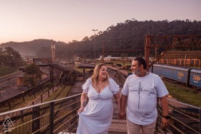 This engaged couple poses for a photo in Vila de Paranapiacaba, Santo André, Brasil, with the pedestrian walkways and bridges over the train rail yard as their backdrop.