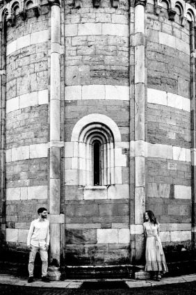 A skilled WPJA photographer captures an engaged couple, standing by an ancient stone structure at Chiesa Madonna del Tiglio in Gravedona, Italy. The timeless setting is rendered in black and white.