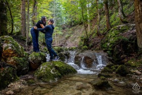 In Villard de Lans, France, nestled on the rocks by a babbling brook under the shade of trees, a couple shares a tender kiss, embodying the natural romance of their surroundings.