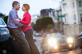 Engaged couple face to face in San Francisco at dusk, man leaning against car, glowing headlights illuminating background street scene.