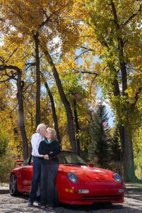 A mature engaged couple snuggles together in a beautiful fall scene in Golden, Colorado, posing with their red sportscar to showcase their personality and shared interests outdoors.