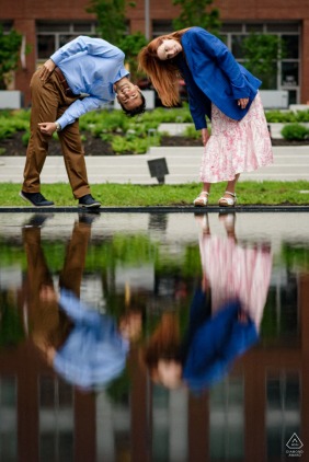 An engaged couple leans over playfully to view their upside-down reflections in a still water pool, creating a unique and artistic engagement portrait in the city of Montreal, Quebec.